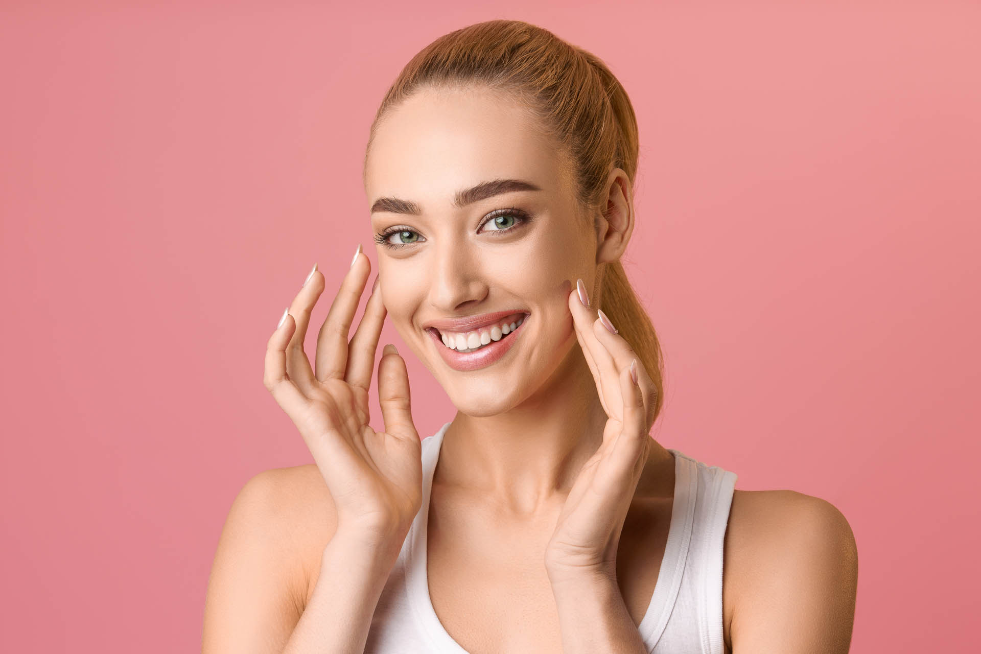 Woman with healthy skin touching her face and smiling to camera, pink background